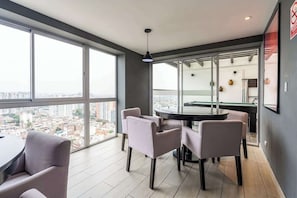 A modern living room with a round table, four chairs, large windows, and a city view. - Geko | Functional Loft next to Malecón w/Pool (Barranco)