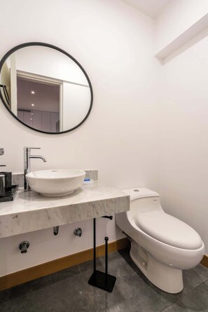 A modern bathroom with a round mirror, a white sink, a chrome faucet, a toilet, and a black floor stand. - Geko | Spectacular 2BR near Malecon w/ balcony (Miraflores)