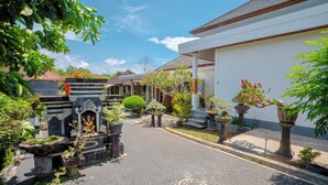 A courtyard with a pathway leading to a building with a white facade and a tiled roof. There are potted plants and a small shrine with a statue. - Moringa Pandawa Guest House (Kutuh)