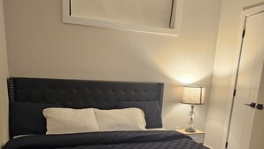 A bedroom with a large bed, a nightstand with a lamp, and a door. - Cozy on 516! Cozy & Comfy 1 bedrm basement suite, Close to Calgary Int'l Airport (Airdrie)