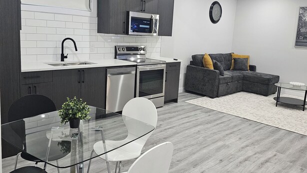 A modern kitchen with white cabinets, a microwave, and a dishwasher. There is a dining area with a glass table and chairs, and a gray sofa in the living room. - Cozy on 516! Cozy & Comfy 1 bedrm basement suite, Close to Calgary Int'l Airport (Airdrie)