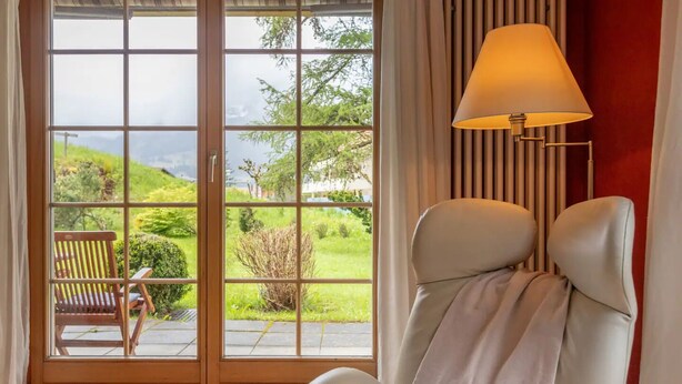 A room with a large sliding glass door leading to a garden, a white chair, and a floor lamp. - Spacious Apartment • Ski-in, Ski-out • Terrace (Lauterbrunnen)