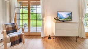 Ein Zimmer mit Holzboden, einem Fernseher auf einem weißen Schrank, einem Sessel mit karierter Decke und Blick auf eine grüne Landschaft durch eine Glastür.
