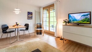 Ein modernes Wohnzimmer mit Holzboden, einem Esstisch mit zwei Stühlen, einem Fernseher auf einem Schrank und einem großen Fenster mit Vorhängen.