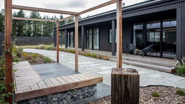 Een modern terras buiten met een houten pergola, een vuurplaats van steen en een zithoek met stoelen.