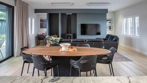 Een moderne woonkamer met een grote televisie, een zwarte leren bank en een eethoek met een houten tafel en zwarte stoelen.