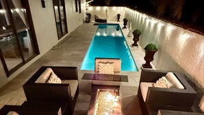 Area piscina con una lunga piscina rettangolare, circondata da divani in vimini scuro con cuscini e un elemento decorativo a forma di focolare al centro.