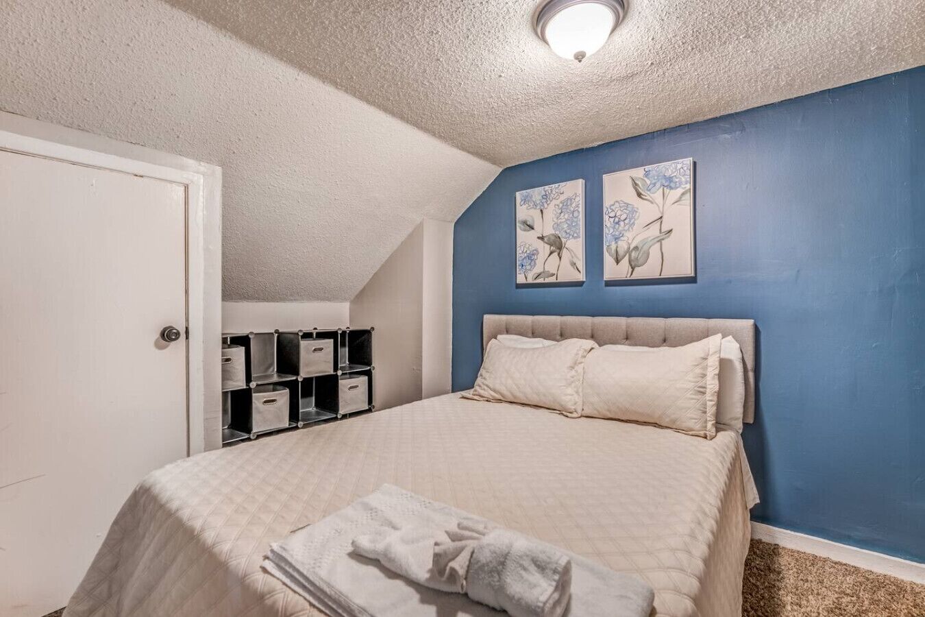 Un dormitorio con una cama, una puerta blanca, una pared azul y dos cuadros con motivos florales.