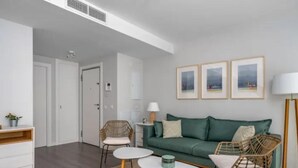 A modern living room with a green sofa, two wicker chairs, a white coffee table, and a floor lamp. There are framed pictures on the wall and a white cabinet with a plant. - H2M Pacificio Seaview & Terrace & Common Pool (Malaga)