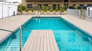 A rectangular swimming pool with a wooden deck in the center, surrounded by potted plants and a modern building with balconies in the background. - H2M Pacificio Seaview & Terrace & Common Pool (Malaga)