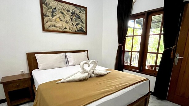 A neatly made bed with a brown blanket and white pillows, a small bedside table, a painting on the wall, and a window with curtains. - Adhya Guest House (Mataram)