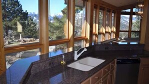 Une cuisine dotée de plans de travail en granit, d’un évier et de meubles en bois. De grandes fenêtres offrent une vue sur les arbres et les montagnes.