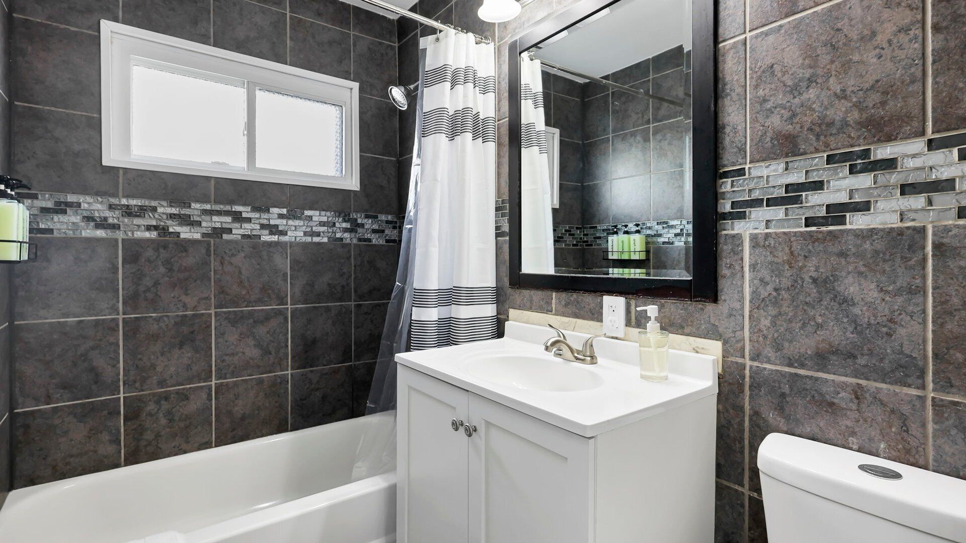 A modern bathroom with a bathtub, a white sink, and a toilet. There is a window above the sink and a shower curtain with horizontal stripes.