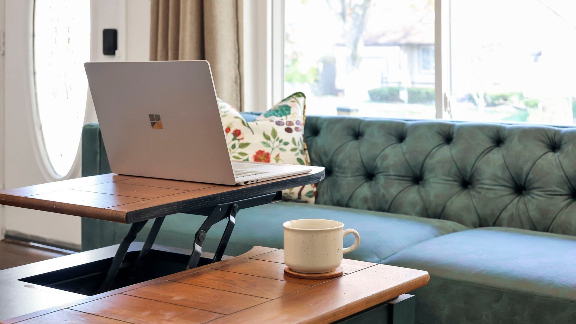 A laptop on a wooden table with a white mug beside it, positioned in front of a green sofa with a floral cushion.