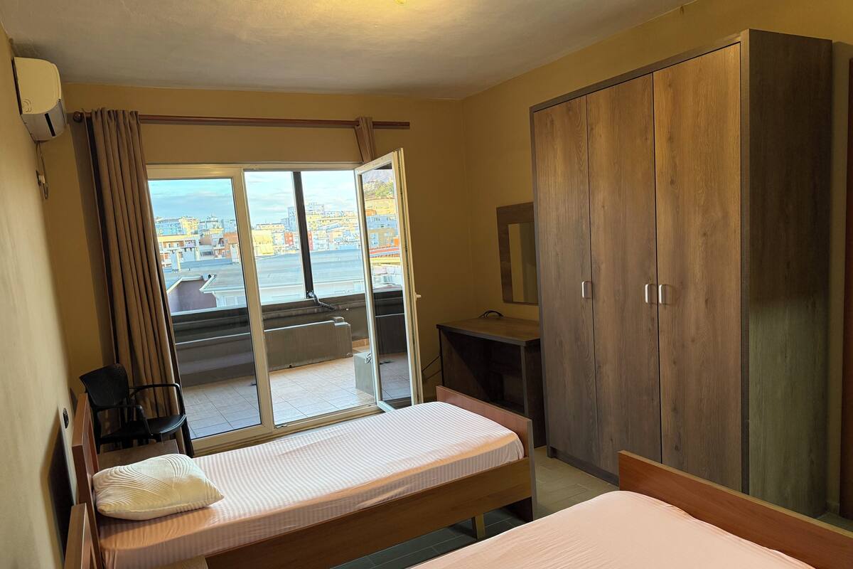 A bedroom with a bed, a desk, a chair, a wardrobe, and a view of the city through large windows.