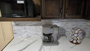 Una cocina con microondas negro, una cafetera Keurig y un cesto de vasos de Dunkin' Donuts.