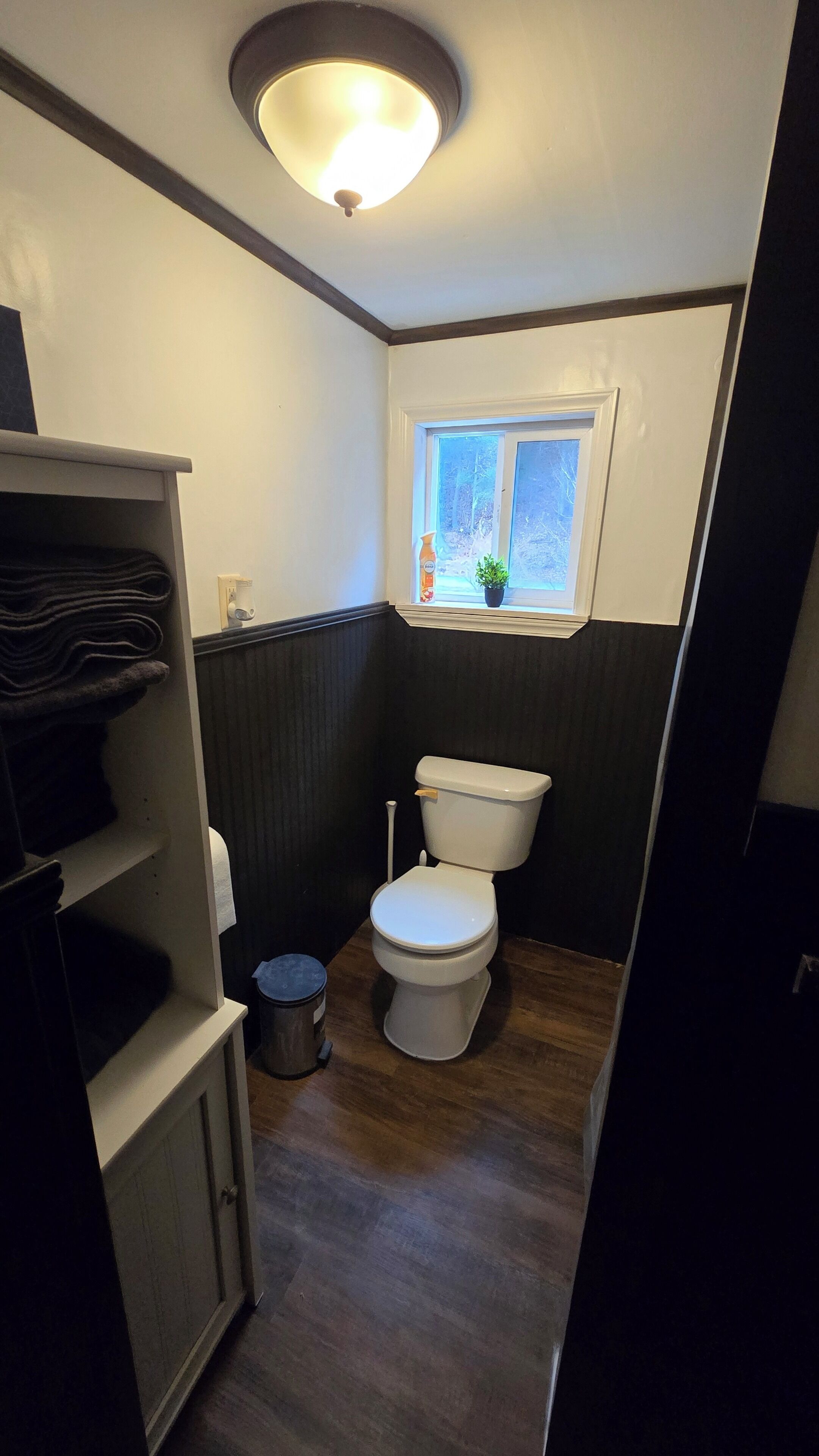 Un baño pequeño con inodoro, un estante con toallas dobladas, un cubo de basura y una ventana con una planta.