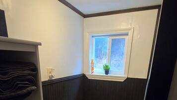 Un baño pequeño con inodoro, un estante con toallas dobladas, un cubo de basura y una ventana con una planta.