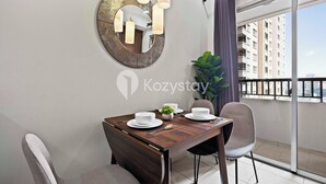Un coin repas avec une table en bois, deux chaises, un miroir rond et une vue sur un balcon agrémenté d’une plante et d’un bâtiment en arrière-plan.