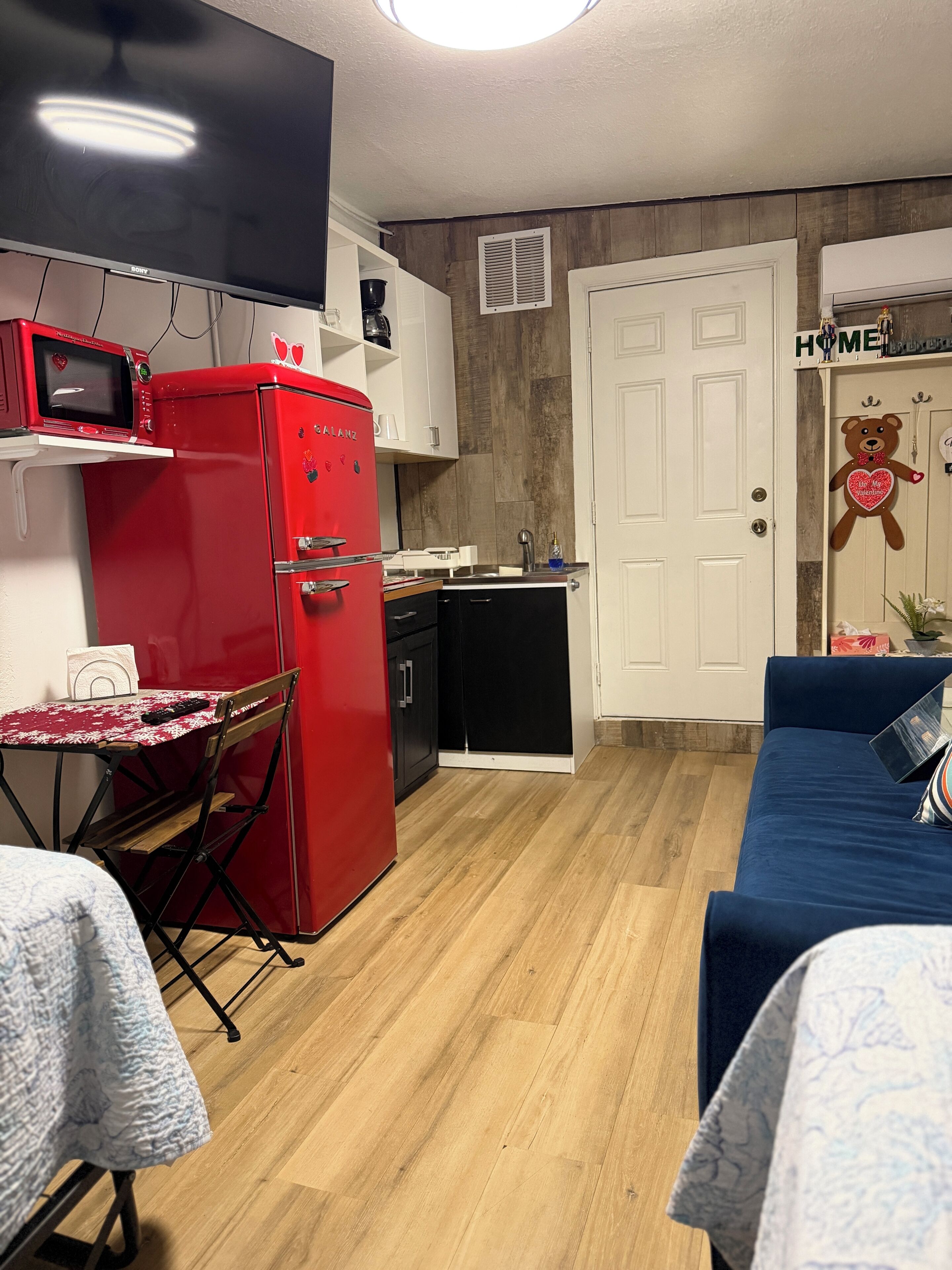 A compact kitchen with a red refrigerator, a microwave, and a sink. There's a dining area with a small table and chairs. The room has wooden flooring and a white door with a teddy bear decoration.
