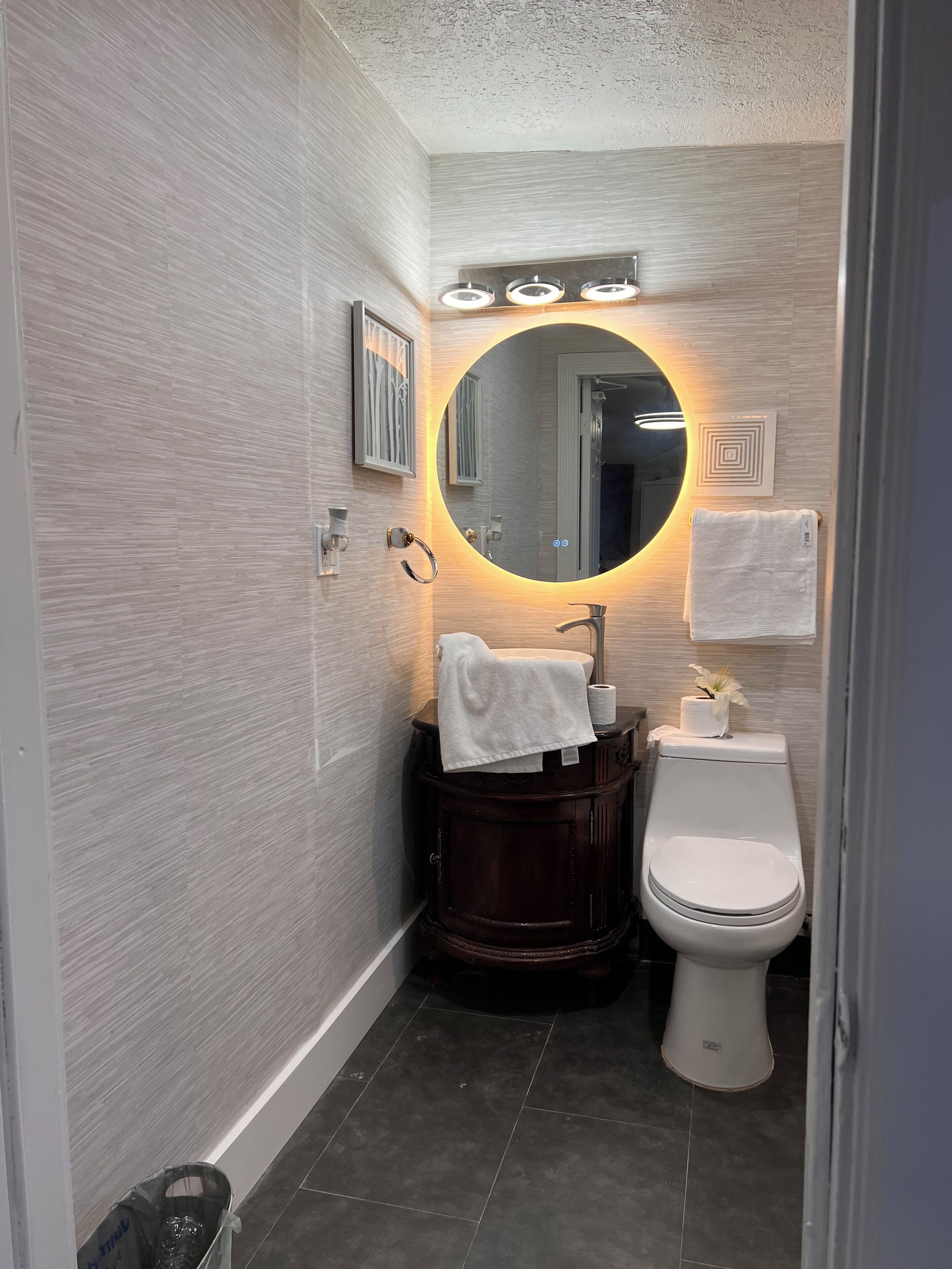 Ein modernes Badezimmer mit einem runden Spiegel, einem dunklen Holzwaschtisch, einer weißen Toilette und einem Handtuch an der Wand.