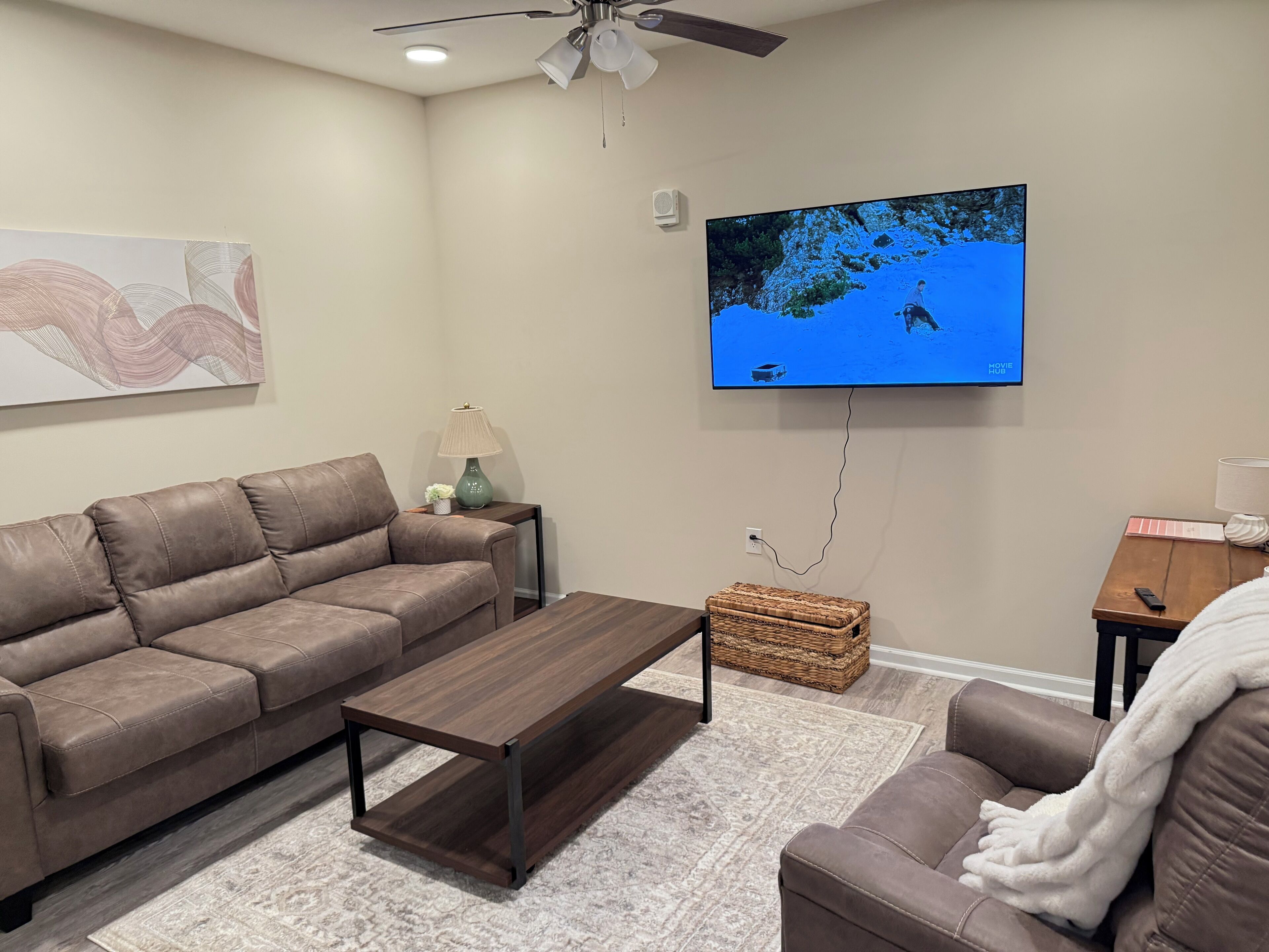 Una sala de estar con un sofá de cuero marrón, una mesa de centro de madera, un televisor montado en la pared y un ventilador de techo.