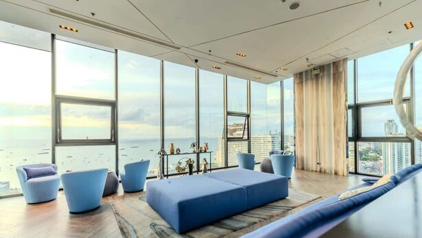 A modern living room with large windows, a blue sofa, and a view of the city. - The Base Pattaya | Top Location & Utility Include (Bang Lamung, Muang Pattaya)