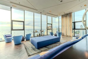 A modern living room with large windows, a blue sofa, and a view of the city. - The Base Pattaya | Top Location & Utility Include (Bang Lamung, Muang Pattaya)