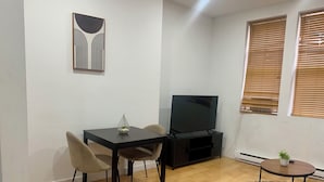 Ein Wohnzimmer mit Holzboden, einem Esstisch mit Stühlen, einem Fernseher und einem kleinen Tisch mit einer Pflanze.