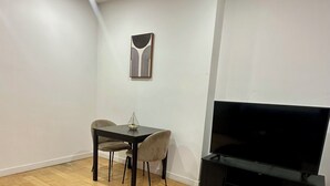 Ein modernes Wohnzimmer mit Holzboden, einem runden Couchtisch mit Pflanze, einem Esstisch mit Stühlen, einem Flachbildfernseher auf einem Schrank und einer Deckenleuchte.