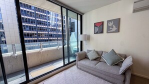 A modern living room with a large sliding glass door leading to a balcony, a grey sofa with cushions, a floor lamp, and two framed artworks on the wall. - Modern CBD Apartment | Pool, Sauna & Laneway Dining (Melbourne)