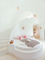 Un dormitorio moderno con decoración blanca, una cama redonda, un pequeño rincón de descanso y una escalera que lleva a un nivel superior.