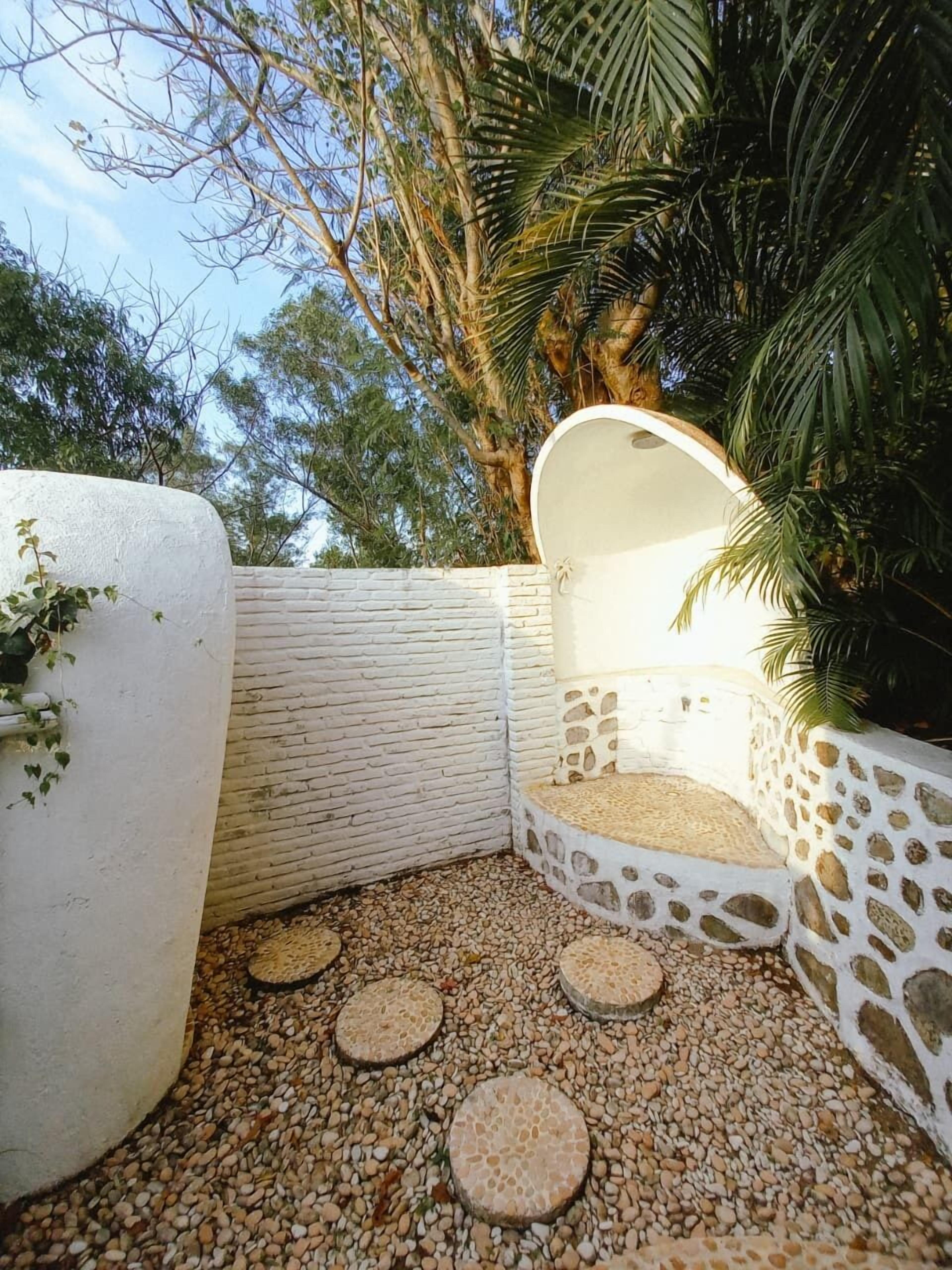 Un jardín en esquina con un muro blanco, un banco de piedra y una palmera.