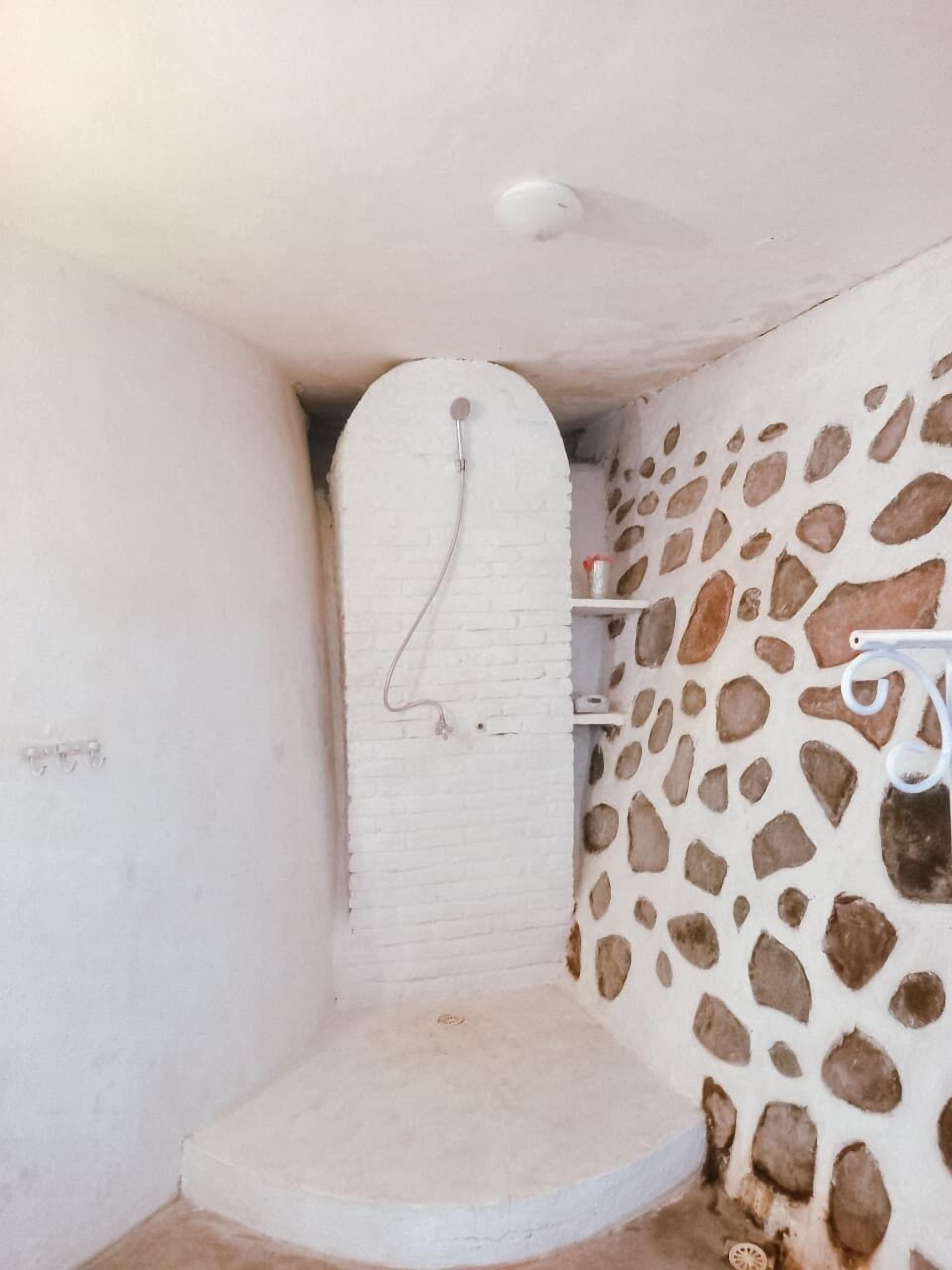 Un espacio de ducha pequeño y cerrado, con un muro de ladrillo blanco y otro con textura de piedra.