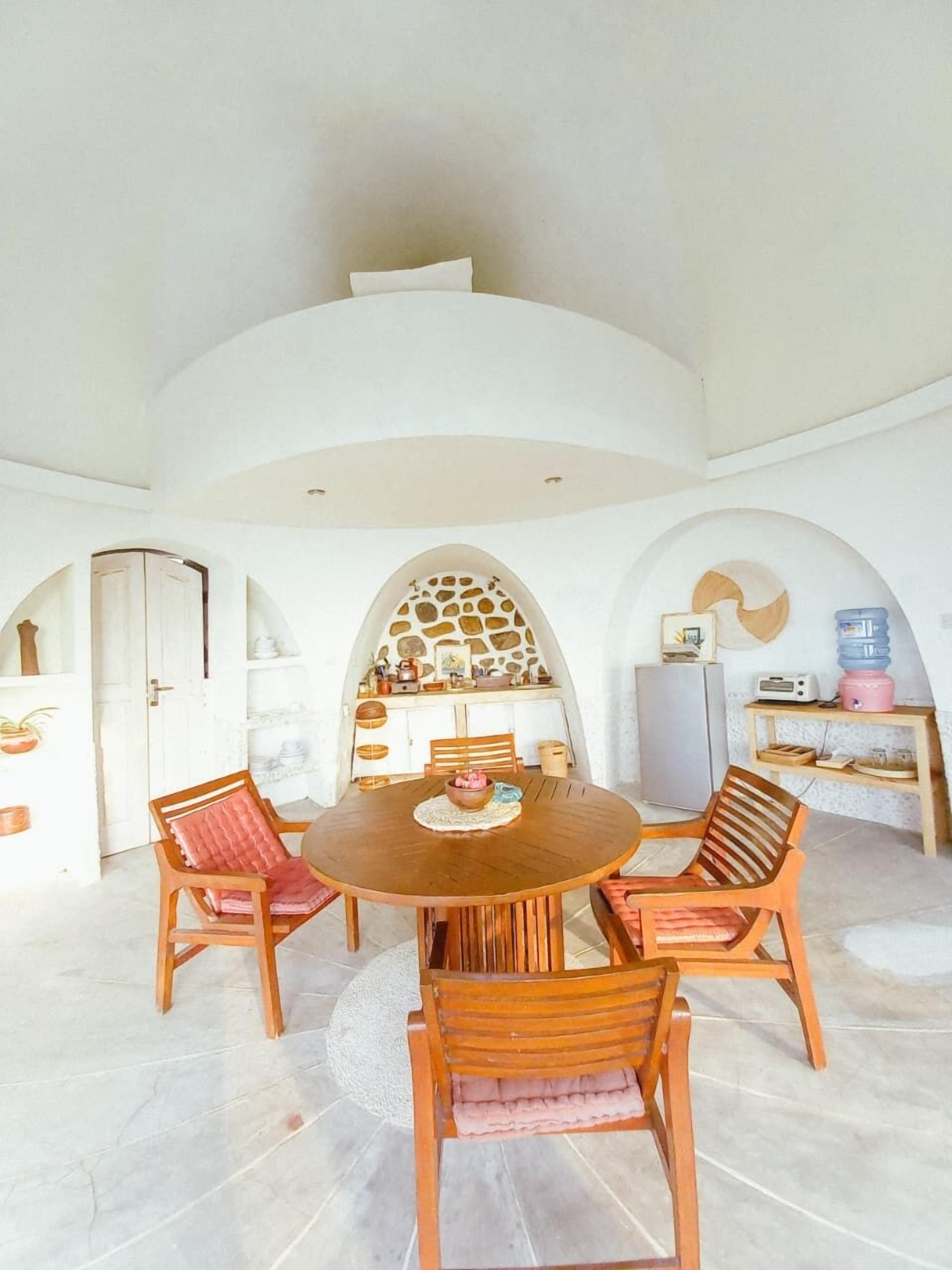 Un coin repas comprenant une table ronde en bois et quatre chaises, un plafond en arc de cercle, ainsi qu’un espace cuisine équipé d’un réfrigérateur et d’un distributeur d’eau.