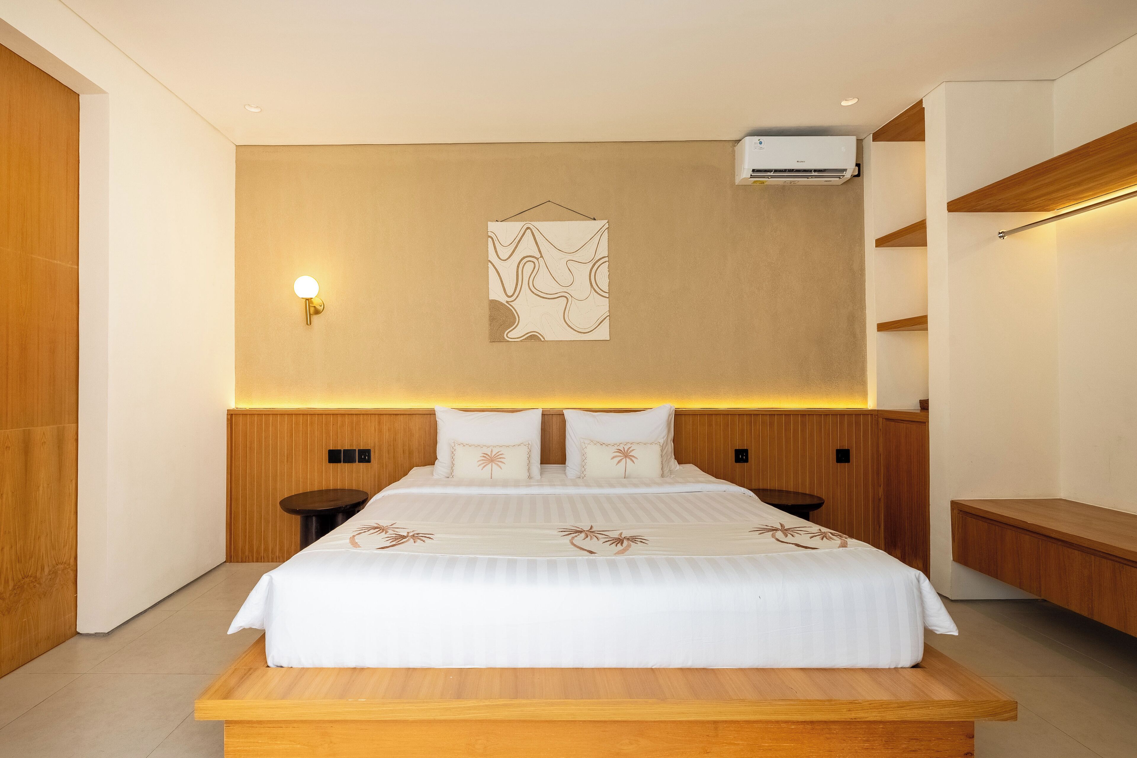 Ett modernt hotellrum med en stor säng, ett träbeklätt sänggavel, sängbord, en väggmonterad luftkonditioneringsanläggning och ett konstverk på väggen.