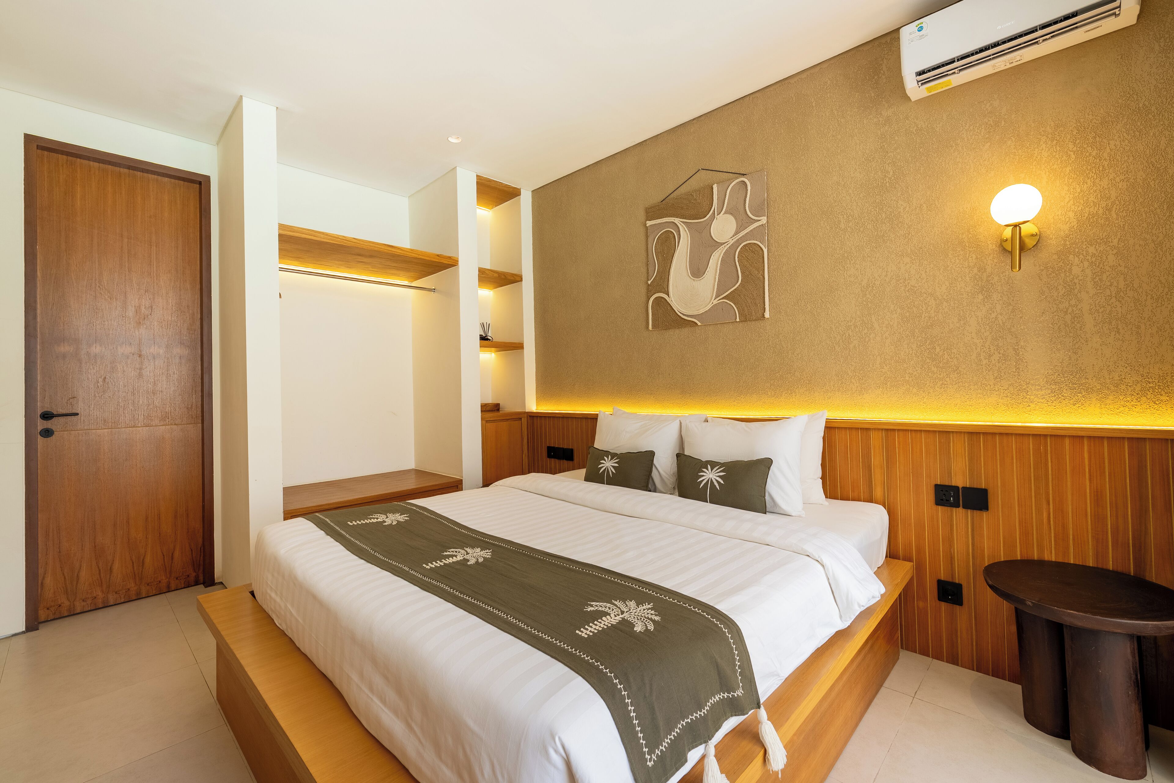 A modern hotel room with a large bed, a bedside table, a wall-mounted air conditioner, and a decorative wall piece.