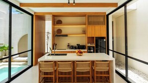 A modern kitchen with a central island, bar stools, and a view of a swimming pool. - Villa Thre by Pertama Stay (Canggu)