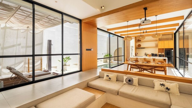 A modern living room with a large window, a sofa, and a dining area with a table and chairs. - Villa Thre by Pertama Stay (Canggu)