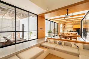 A modern living room with a large window, a sofa, and a dining area with a table and chairs. - Villa Thre by Pertama Stay (Canggu)