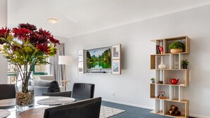 A modern living room with a glass dining table, comfortable seating, a mounted TV, and a wooden shelving unit with decorative items. - Family Unit With Balcony, Free Trams to CBD Sights (Melbourne)