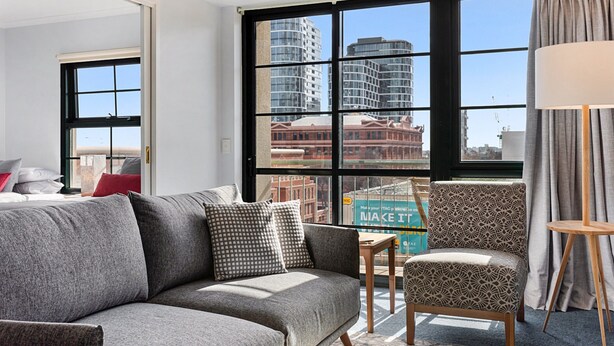 A modern living room with a grey sofa, patterned armchair, and a view of city buildings through large windows. - Family Unit With Balcony, Free Trams to CBD Sights (Melbourne)