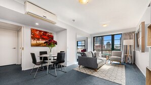 A modern living room with a dining area, a grey sofa, a coffee table, and a large window offering a city view. - Family Unit With Balcony, Free Trams to CBD Sights (Melbourne)