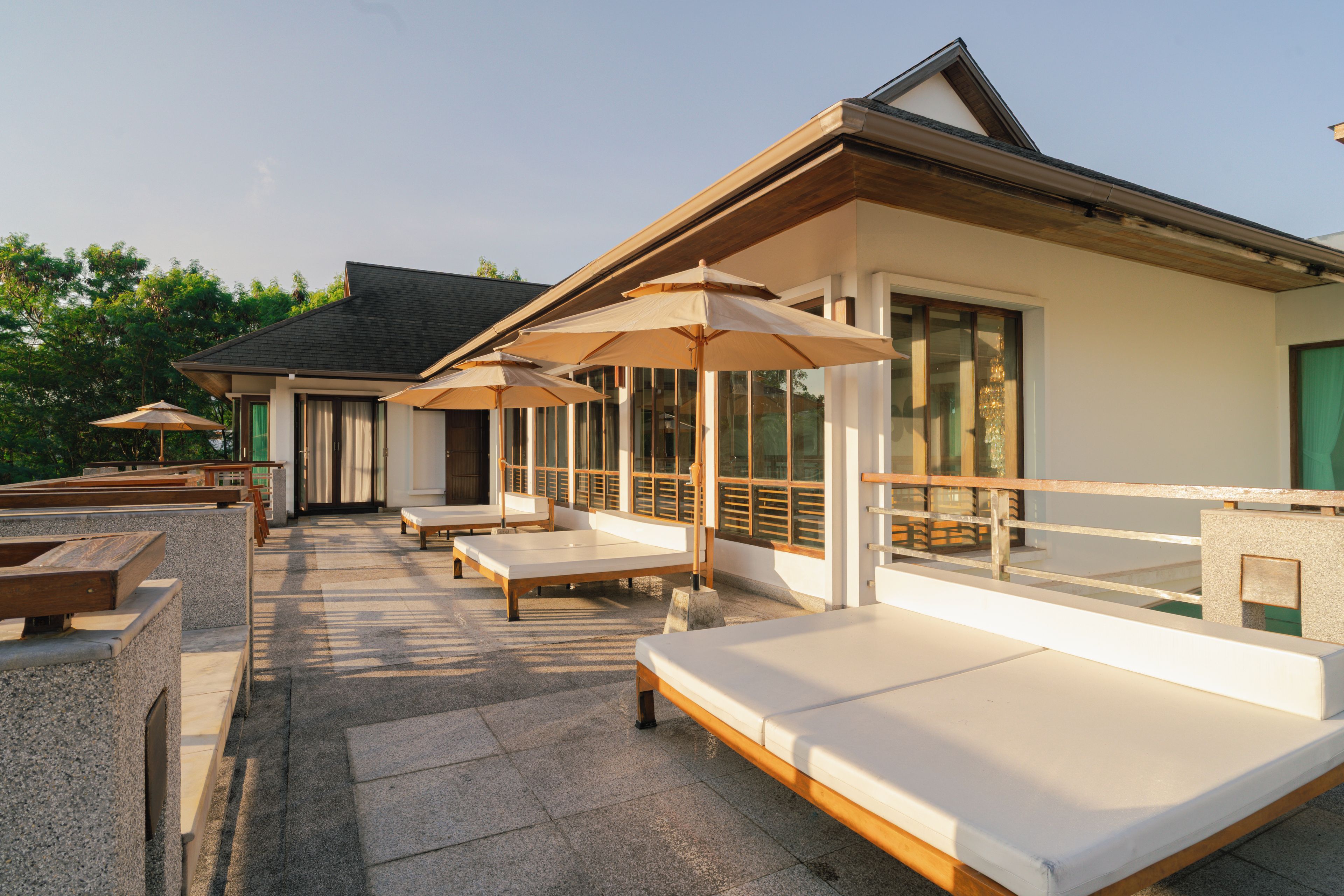 Un área de estar moderna al aire libre con sillones reclinables y sombrillas, piso de piedra y un edificio con grandes ventanales.