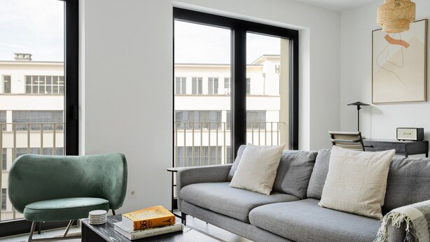 Una sala moderna con un sofá gris, un sillón verde y una mesa de centro. Hay ventanas amplias con vistas a edificios.