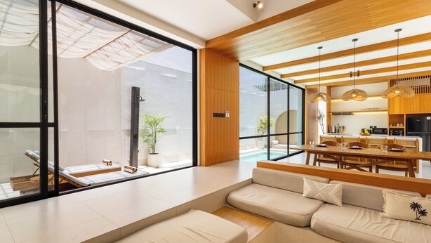 A modern living room with a large window, a sofa, a dining area, and a kitchen visible through the glass wall. - Villa Tin by Pertama Stay (Canggu)