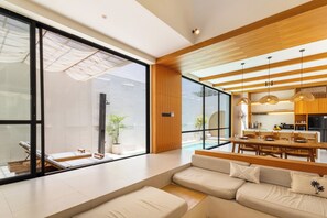 A modern living room with a large window, a sofa, a dining area, and a kitchen visible through the glass wall. - Villa Tin by Pertama Stay (Canggu)