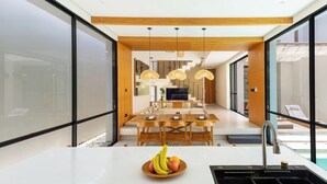 A modern kitchen with a central island, a dining area with a wooden table and chairs, and a view of a pool and lounge area. - Villa Tin by Pertama Stay (Canggu)