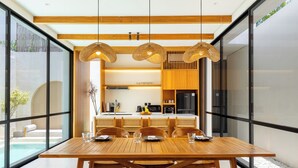 A modern dining area with a wooden table and chairs, large windows, and a kitchen in the background. - Villa Tin by Pertama Stay (Canggu)