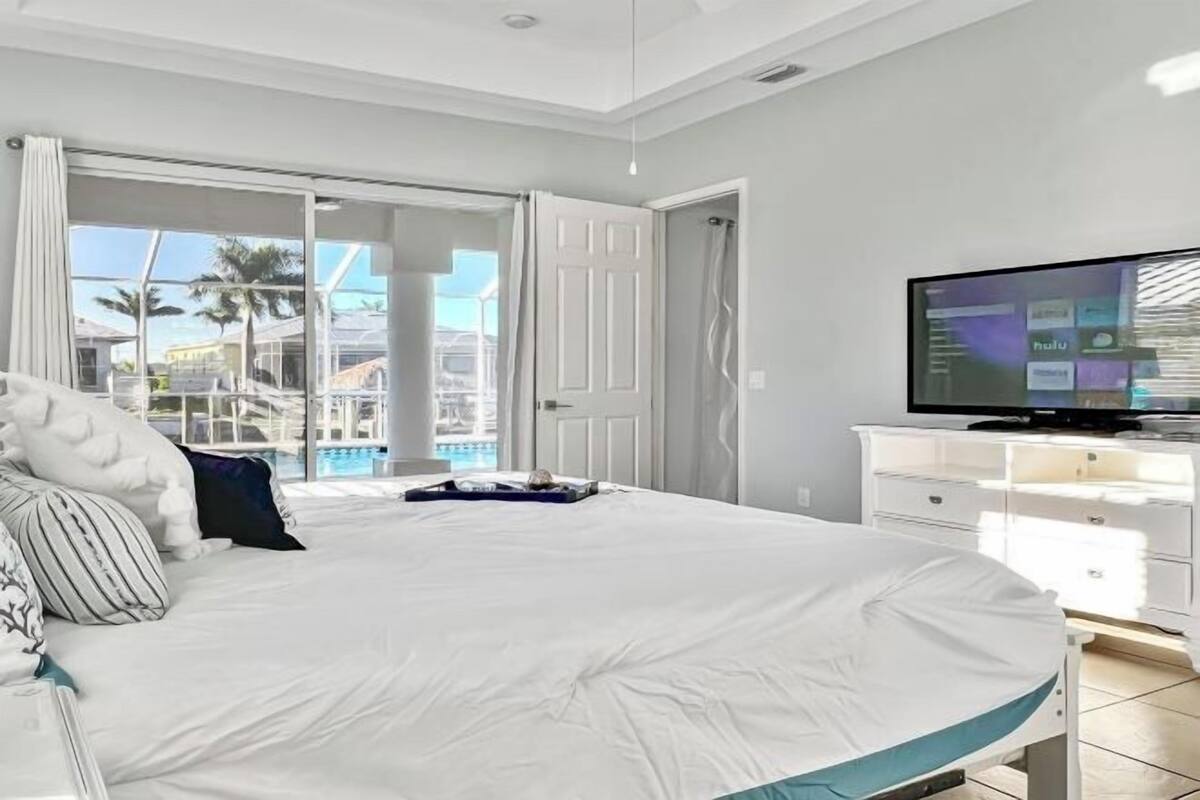 A bedroom with a bed, a television, a white dresser, and a sliding glass door leading to a balcony.